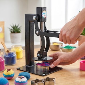 May include: A black metal bath bomb press machine with a vertical lever and a circular pressing plate. The machine features a small circular logo with the text Mooy Bath. It is positioned on a wooden surface alongside various colorful powders.