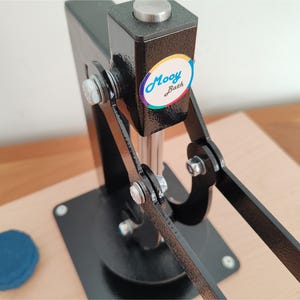 Op de afbeelding: Een zwarte metalen pers met een zilveren handvat en voet, gebruikt voor knutselen. De pers heeft een kleurrijke "Mooy Bath" sticker. Een klein, blauw item ligt op het houten oppervlak.