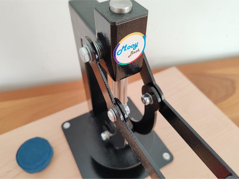 Op de afbeelding: Een zwarte metalen pers met een zilveren handvat en voet, gebruikt voor knutselen. De pers heeft een kleurrijke "Mooy Bath" sticker. Een klein, blauw item ligt op het houten oppervlak.