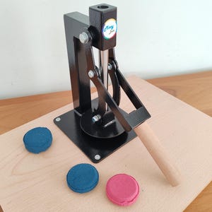 Op de afbeelding: Een zwarte metalen macaronpers met een houten handvat en een ronde basis, geplaatst op een licht houten oppervlak. Drie macarons in blauw en roze liggen in de buurt van de pers. De pers heeft de tekst "Moony Sue".