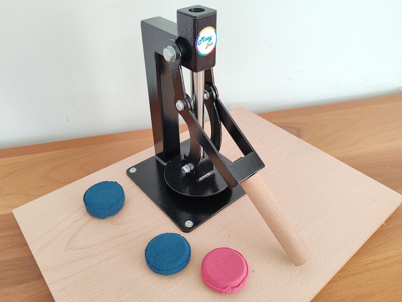 Op de afbeelding: Een zwarte metalen macaronpers met een houten handvat en een ronde basis, geplaatst op een licht houten oppervlak. Drie macarons in blauw en roze liggen in de buurt van de pers. De pers heeft de tekst "Moony Sue".