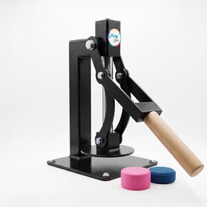 May include: A black metal button press machine with a wooden handle and a vertical lever mechanism. The machine features a small circular logo with the text Mooy Jack. Two small circular plastic components, one pink and one blue, sit in front.