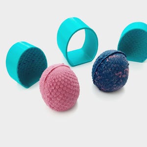 Op de afbeelding: Een bovenaanzicht toont een verzameling badbruisballen en turquoise ringvormige objecten. Twee roze badbruisballen met een textuuroppervlak liggen naast een blauwe badbruisbal. De turquoise ringen zijn rond de badbruisballen geplaatst.