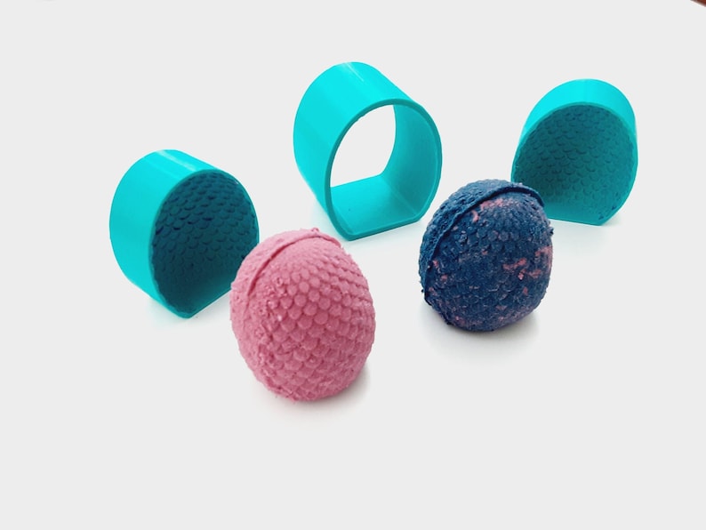 Op de afbeelding: Een bovenaanzicht toont een verzameling badbruisballen en turquoise ringvormige objecten. Twee roze badbruisballen met een textuuroppervlak liggen naast een blauwe badbruisbal. De turquoise ringen zijn rond de badbruisballen geplaatst.