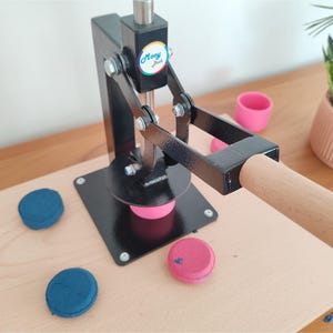 Op de afbeelding: Een zwarte knopenmaker met een houten handvat en basis. De machine drukt op een roze knop. Twee blauwe knopen en een roze knop liggen op het houten oppervlak. Een klein roze bekertje en een plant in een pot staan op de achtergrond.