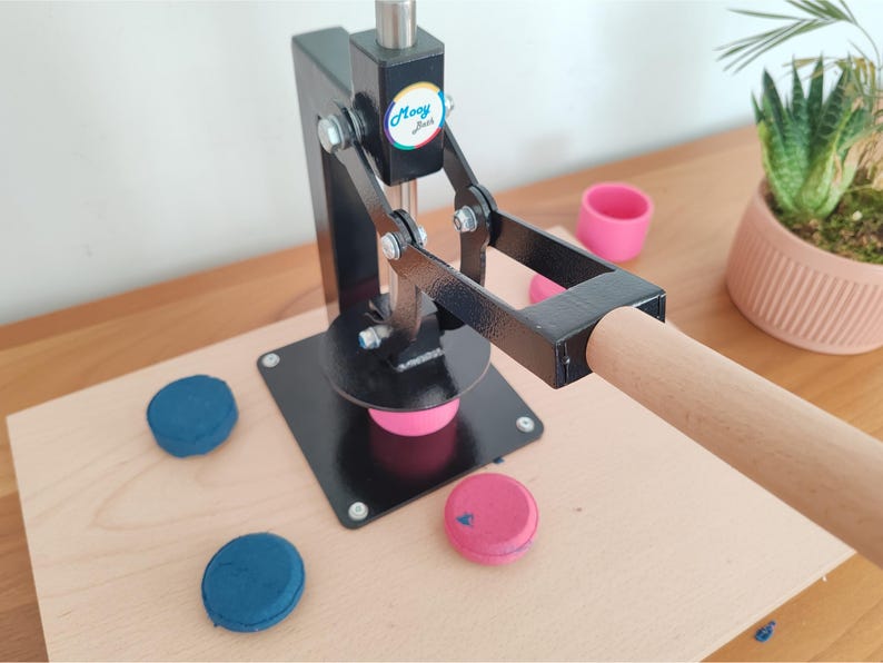 Op de afbeelding: Een zwarte knopenmaker met een houten handvat en basis. De machine drukt op een roze knop. Twee blauwe knopen en een roze knop liggen op het houten oppervlak. Een klein roze bekertje en een plant in een pot staan op de achtergrond.