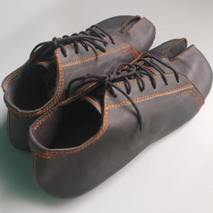 May include: A pair of black leather split-toe shoes with orange stitching and black laces. The shoes have a unique design, with a separate compartment for the big toe. The shoes are designed for comfort and flexibility.