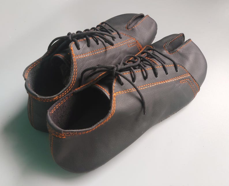 May include: A pair of black leather split-toe shoes with orange stitching and black laces. The shoes have a unique design, with a separate compartment for the big toe. The shoes are designed for comfort and flexibility.