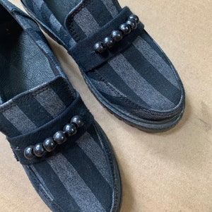 Peut inclure: Paire de mocassins rayés noirs et gris avec un ornement de perles noires. Les chaussures ont un design classique à enfiler avec un bout arrondi et un petit talon. Le tissu a un éclat subtil.