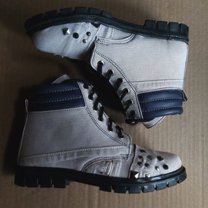 Puede incluir: Un par de botas de lona grises con suelas negras y detalles azules. Las botas tienen tachuelas plateadas en la punta y los lados.