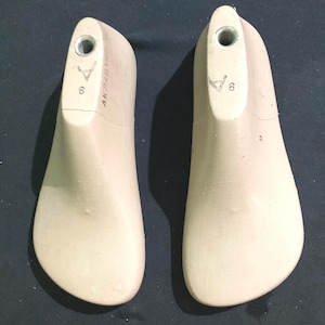 May include: Two beige shoe lasts, size 6, made of a solid material. Each has a metal insert at the top and is marked with the text "AK240306". The lasts are designed for shoemaking and are placed on a dark surface.