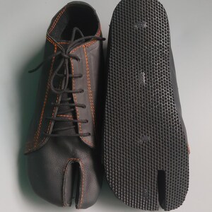 May include: Black split-toe shoes with orange stitching and black laces. The sole is textured with a split toe design. The shoes are made of a leather-like material and are designed for comfort and flexibility.