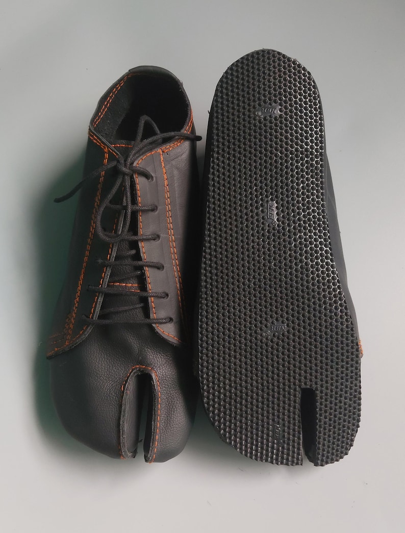 May include: Black split-toe shoes with orange stitching and black laces. The sole is textured with a split toe design. The shoes are made of a leather-like material and are designed for comfort and flexibility.