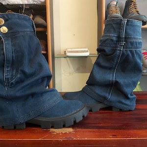 May include: A pair of blue denim boots with a folded cuff. The boots have a chunky black rubber sole and are sitting on a wooden surface.
