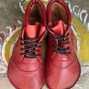 Op de afbeelding: Een paar rode leren schoenen met zwarte veters. De schoenen hebben een afgeronde neus en een gestikte naad rond de rand.