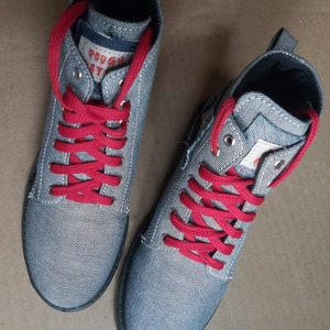 Può includere: Un paio di sneakers alte in denim grigio con lacci rossi. Le sneakers hanno un'etichetta bianca sul lato che dice "Rough Trade".