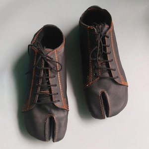 May include: A pair of black leather split-toe shoes with orange stitching and black laces. The shoes have a unique design with a separate compartment for the big toe, and are likely designed for comfort and flexibility.