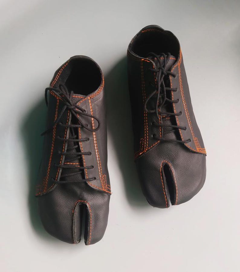 May include: A pair of black leather split-toe shoes with orange stitching and black laces. The shoes have a unique design with a separate compartment for the big toe, and are likely designed for comfort and flexibility.
