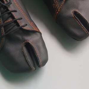 May include: A pair of black leather split-toe shoes with orange stitching and black laces. The shoes have a unique design with a separate compartment for the big toe. The shoes are on a white surface.