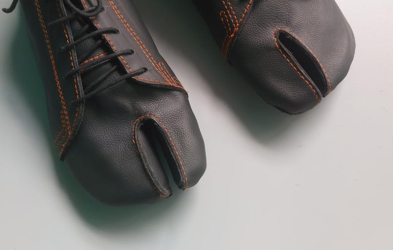 May include: A pair of black leather split-toe shoes with orange stitching and black laces. The shoes have a unique design with a separate compartment for the big toe. The shoes are on a white surface.