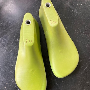 Könnte beinhalten: Zwei hellgrüne Schuhleisten, Größe 11 1/2, werden gezeigt. Jede Leiste hat eine abgerundete Spitze und ein Loch an der Ferse. Die Leisten bestehen aus einem festen Material und haben eine glatte, leicht strukturierte Oberfläche. Die Nummer 11 1/2 ist auf der Ferse jeder Leiste aufgedruckt.