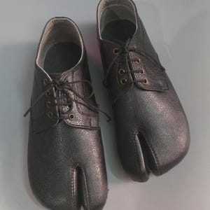 Puede incluir: Un par de zapatos negros de cuero con punta dividida y cordones. Los zapatos tienen una punta redonda y un tacón bajo.