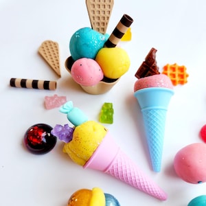 Könnte beinhalten: Eine Auswahl an bunten, kleinen Eiswaffeln und Eisbechern. Das Bild zeigt Eiscremekugeln in Blau, Gelb und Rosa, mit Waffel- und Schokoladenakzenten. Andere Süßigkeiten sind um das Eis herum verstreut.