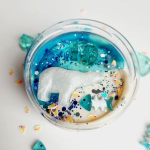 Könnte beinhalten: Ein klarer Behälter gefüllt mit einem geschichteten Wachsschmelz. Die obere Schicht ist blau mit Glitzer und einer klaren Eiswürfelform. Ein weißer Eisbär und fischförmige Wachsschmelze befinden sich im Behälter.