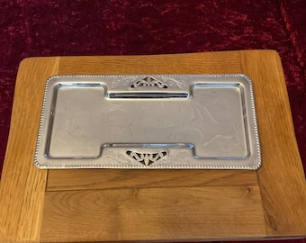 Puede incluir: Una bandeja rectangular plateada con bordes decorativos y detalles ornamentados, colocada sobre una superficie de madera. La bandeja tiene dos áreas empotradas y está diseñada para servir alimentos o exhibir artículos.