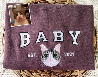 Puede incluir: Sudadera color burdeos con el texto bordado "BABY EST. 2021" y una imagen de gato bordada.