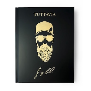 Könnte beinhalten: Schwarzes Notizbuch mit goldfarbenem Schriftzug und einer goldenen Illustration eines Mannes mit Mütze und Brille. Das Wort "TUTTAVIA" steht oben, mit einer Unterschrift unten.