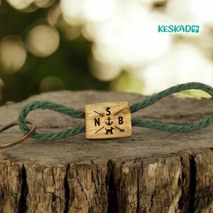 May include: A wooden keychain with a green rope loop and a brass key ring. The wooden block is engraved with the initials "S", "N", and "B", along with an anchor and a dog silhouette. The keychain is resting on a wooden surface.