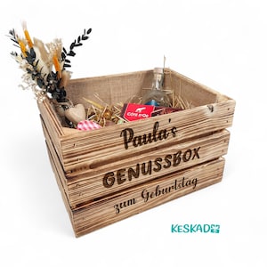 Könnte beinhalten: Eine Holzkiste mit der Gravur "Paula's GENUSSBOX zum Geburtstag". Die Kiste ist mit einer Flasche, Pralinen und dekorativen Blumen gefüllt.