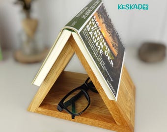 May include: A wooden triangle book stand with a book titled "Das Geheime Leben Der Bäume" resting on top. A pair of black glasses is resting on the bottom shelf of the stand.
