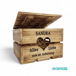 May include: A wooden box with a hinged lid, featuring the name "SANDRA" and the words "Alles Liebe zum 40. Geburtstag" with a heart-shaped cutout. The box is made of light brown wood and contains straw.