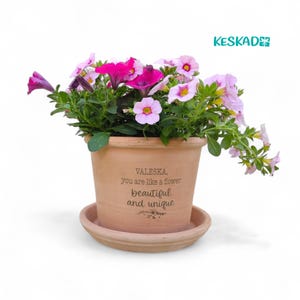 Könnte beinhalten: Ein Terrakotta-Blumentopf gefüllt mit leuchtend rosa und lila Petunien. Der Topf trägt die Aufschrift "VALESKA, you are like a flower beautiful and unique". Der Topf steht auf einer passenden Untertasse.