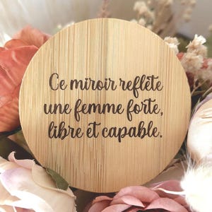 Peut inclure: Un disque rond en bois clair portant l'inscription "Ce miroir reflète une femme forte, libre et capable." en écriture noire. Le disque est entouré de fleurs et de feuillages roses et pêche.