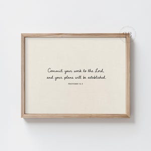 May include: A rectangular wooden frame containing a cream-colored print with the black text: Commit your work to the Lord, and your plans will be established. PROVERBS 16:3. The frame has a natural light brown wood grain finish and a simple profile.