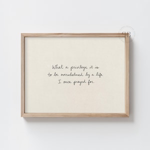 May include: A rectangular wooden frame containing a cream-coloured print with the black handwritten-style text: What a privilege it is to be overwhelmed by a life I once prayed for. The frame has a natural light wood finish and a simple profile.