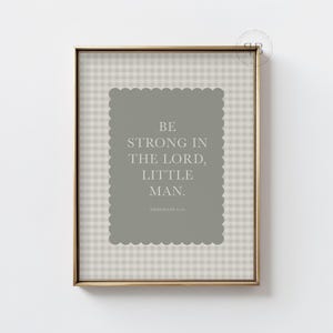 May include: Framed print with the text "BE STRONG IN THE LORD, LITTLE MAN." The text is in white on a gray, scalloped-edge background. The background is a light gray and white checkered pattern. The frame is gold.