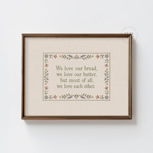 May include: A framed cross-stitch embroidery on light beige fabric featuring the text: We love our bread, we love our butter, but most of all, we love each other. The text is surrounded by a decorative border of small, colorful floral motifs.