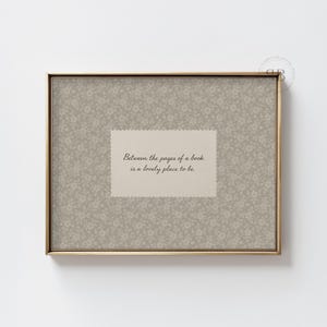 May include: A framed print with a gold-colored frame. The print features a floral pattern background in shades of gray and a rectangular insert with the text: "Between the pages of a book is a lovely place to be."