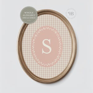 May include: An oval framed print with a pink background and a white letter "S" inside a floral wreath. The background has a gingham pattern. The frame is gold. The text "WHOLE ALPHABET INCLUDED" is in a circle at the top.