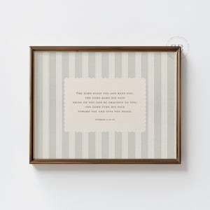 Può includere: Una stampa artistica rettangolare da parete in una cornice color bronzo scuro con uno sfondo a righe verticali grigio chiaro e bianco. Un'etichetta centrale color crema con bordi smerlati riporta il testo: The Lord bless you and keep you; the Lord make his face shine on you and be gracious to you; the Lord turn his face toward you and give you peace. Numbers 6:24-26.