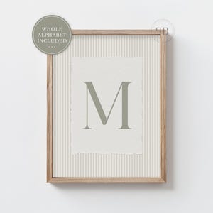 May include: A framed print with the letter "M" in a muted green colour. The letter is on textured, off-white paper with a torn edge, set against a striped background. The frame is light-coloured wood. A circular graphic says "WHOLE ALPHABET INCLUDED".