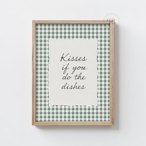 May include: A framed print with a light wood frame. The print features a green and white gingham pattern and a central piece of paper with the text "Kisses if you do the dishes" in black script.