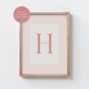 May include: A framed print featuring the letter "H" in a soft pink hue. The letter is centered on a textured white paper, set against a pink and white striped background. The frame is a light wood color, and a circular sticker in the upper left corner reads "WHOLE ALPHABET INCLUDED".
