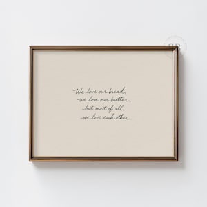 May include: A framed print with a gold-coloured frame. The print features a beige background with black cursive text: "We love our bread, we love our butter, but most of all, we love each other."