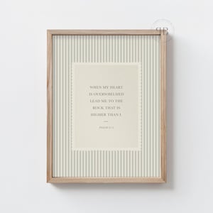 May include: A framed print with a light wood frame. The print has a striped background and a central cream-colored rectangle with text that reads, "WHEN MY HEART IS OVERWHELMED LEAD ME TO THE ROCK THAT IS HIGHER THAN I." Psalm 61:2 is below.