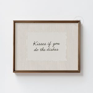 May include: A framed print with the text "Kisses if you do the dishes" in a handwritten font. The frame is a warm brown color, and the background has vertical stripes. The paper has a textured, torn edge.